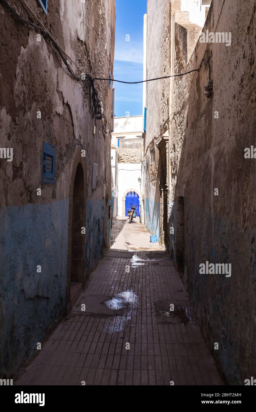 Narrow street full of shadow, with an enlightened white house in the background. Bright blue gate with an arc and a motorbike infront. Essaouira, Moro Stock Photo