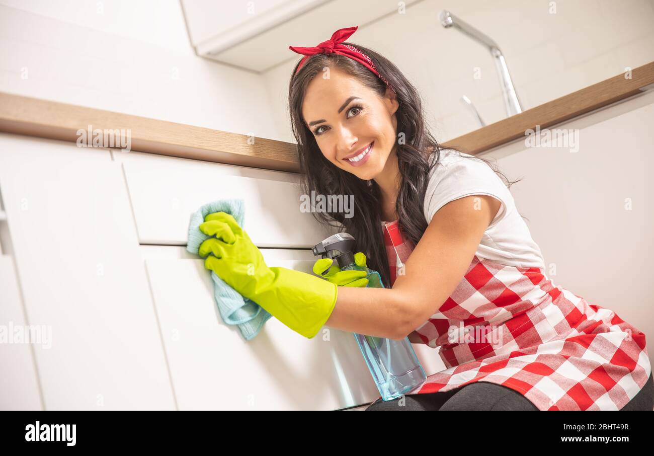 Lovely young housemaid in apron cleaning white kitchen cupboard door with a cloth and detergent in yellow rubber gloves. Stock Photo