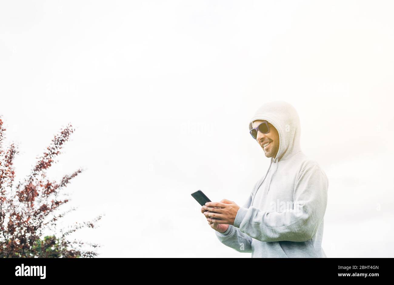 Young man in gray sweatshirt and blue sunglasses holding cell phone in his hands and smiling with trees and a white background Stock Photo
