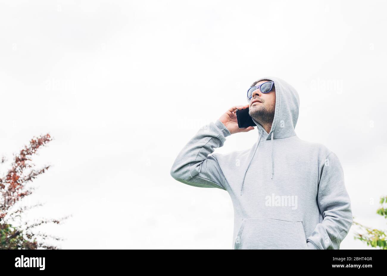 Young man in gray sweatshirt and blue sunglasses holding cell phone in his hands and smiling on a white background Stock Photo