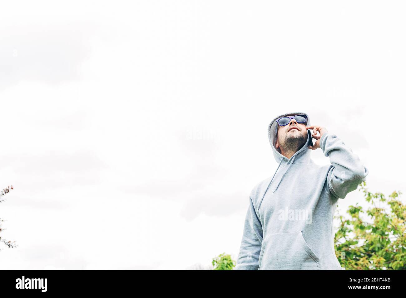 Young man in gray sweatshirt and blue sunglasses holding cell phone in his hands looking up Stock Photo
