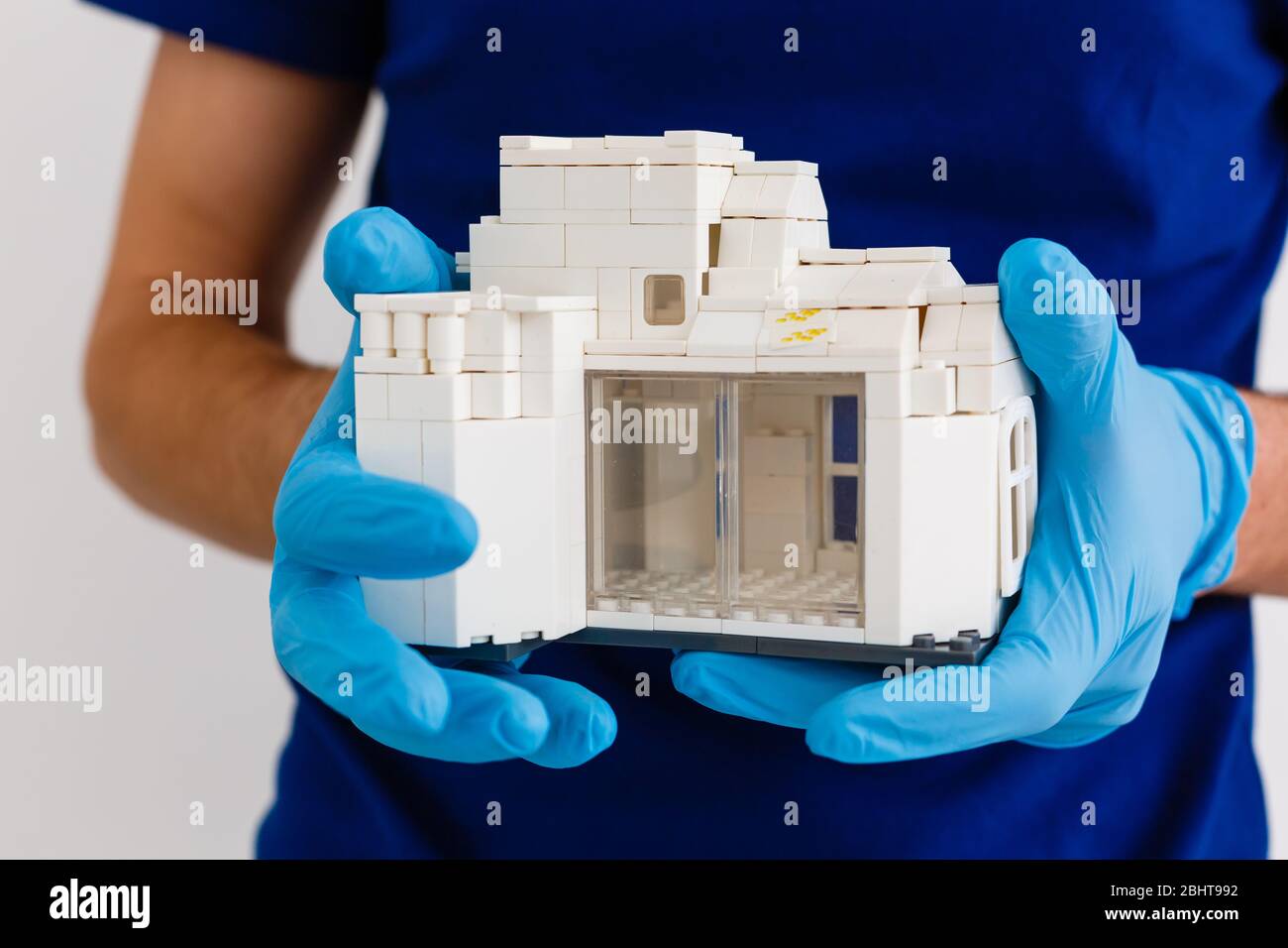 small wooden house in the palm of your hand with a white medical rubber glove, blue background, wooden model of the house, quarantine concept Stock Photo