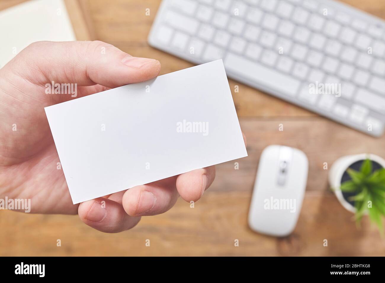 A hand holding white empty business card over wooden table in office. Top view. Stock Photo
