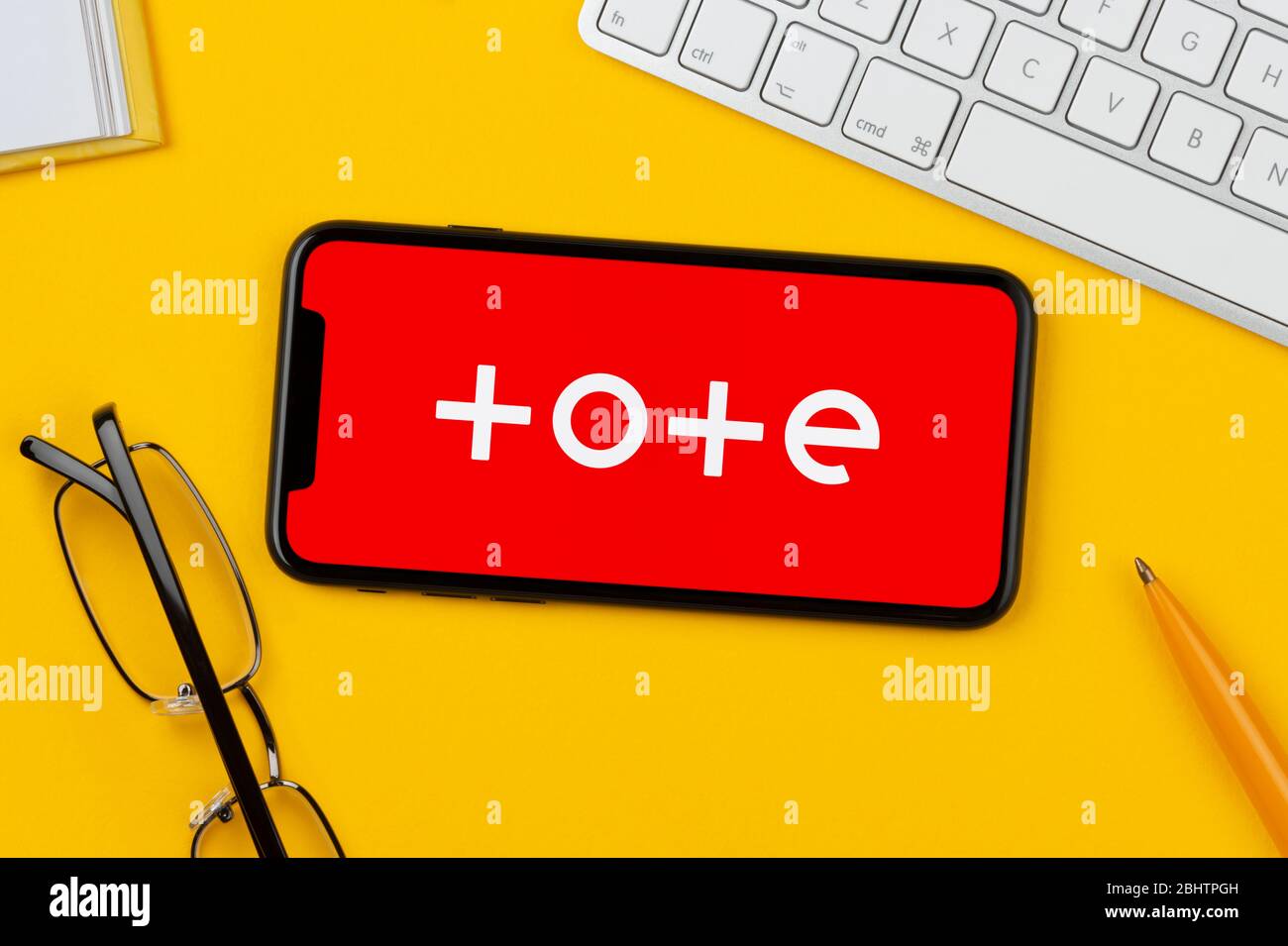 A smartphone showing the Tote logo rests on a yellow background along with a keyboard, glasses, pen and book (Editorial use only). Stock Photo