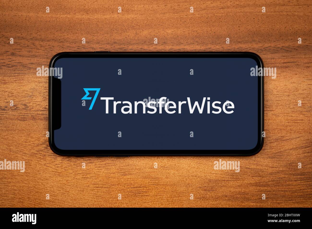 A smartphone showing the Transferwise logo rests on a plain wooden table (Editorial use only). Stock Photo
