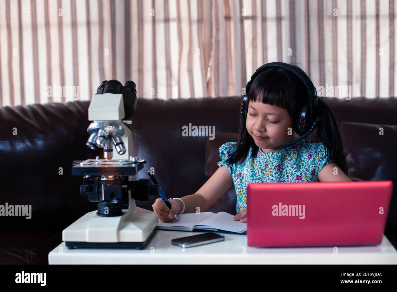 Asian little child  girl wearing headphones learning online by using laptop and microscope at home , distance education Stock Photo