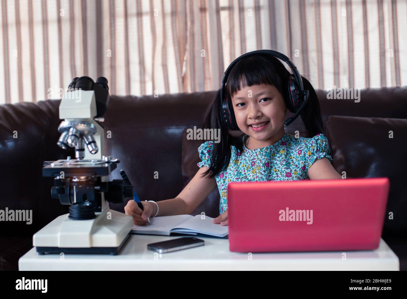 Asian little child  girl wearing headphones learning online by using laptop and microscope at home , distance education Stock Photo
