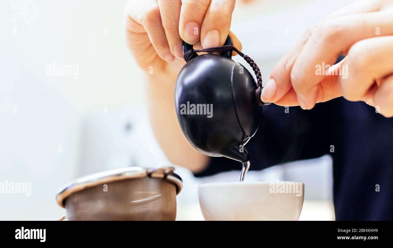 Close-up of hands pouring Chinese green tea from the small ceramic kettle into a small cup with tea strainer in foreground. Stock Photo