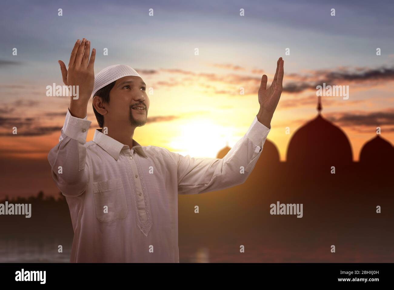 Asian Muslim man standing while raised hands and praying with a sunset sky background Stock Photo