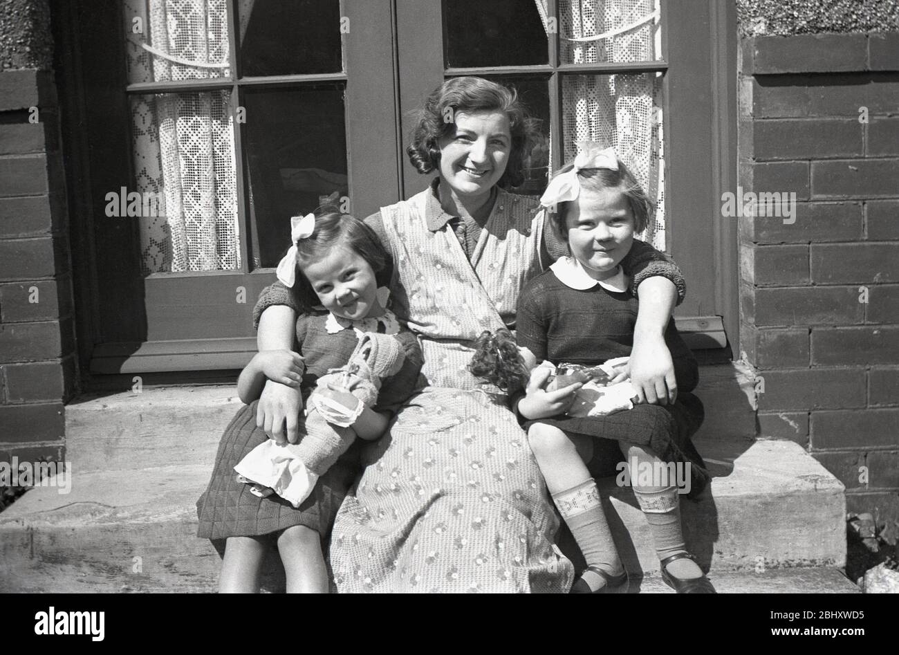 1950s, historical, a mother and housewife wearing a pinafore, sitting on the steps of the back doors of a house with her arms around her two daughters, two sweet little girls with ribbons in their hair and who are both holding their toy dolls, England, UK. Stock Photo