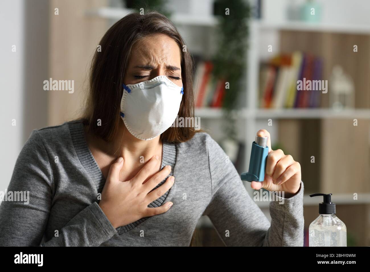 Woman with protective mask suffocating due covid-19 holds inhaler sitting on a desk at night Stock Photo