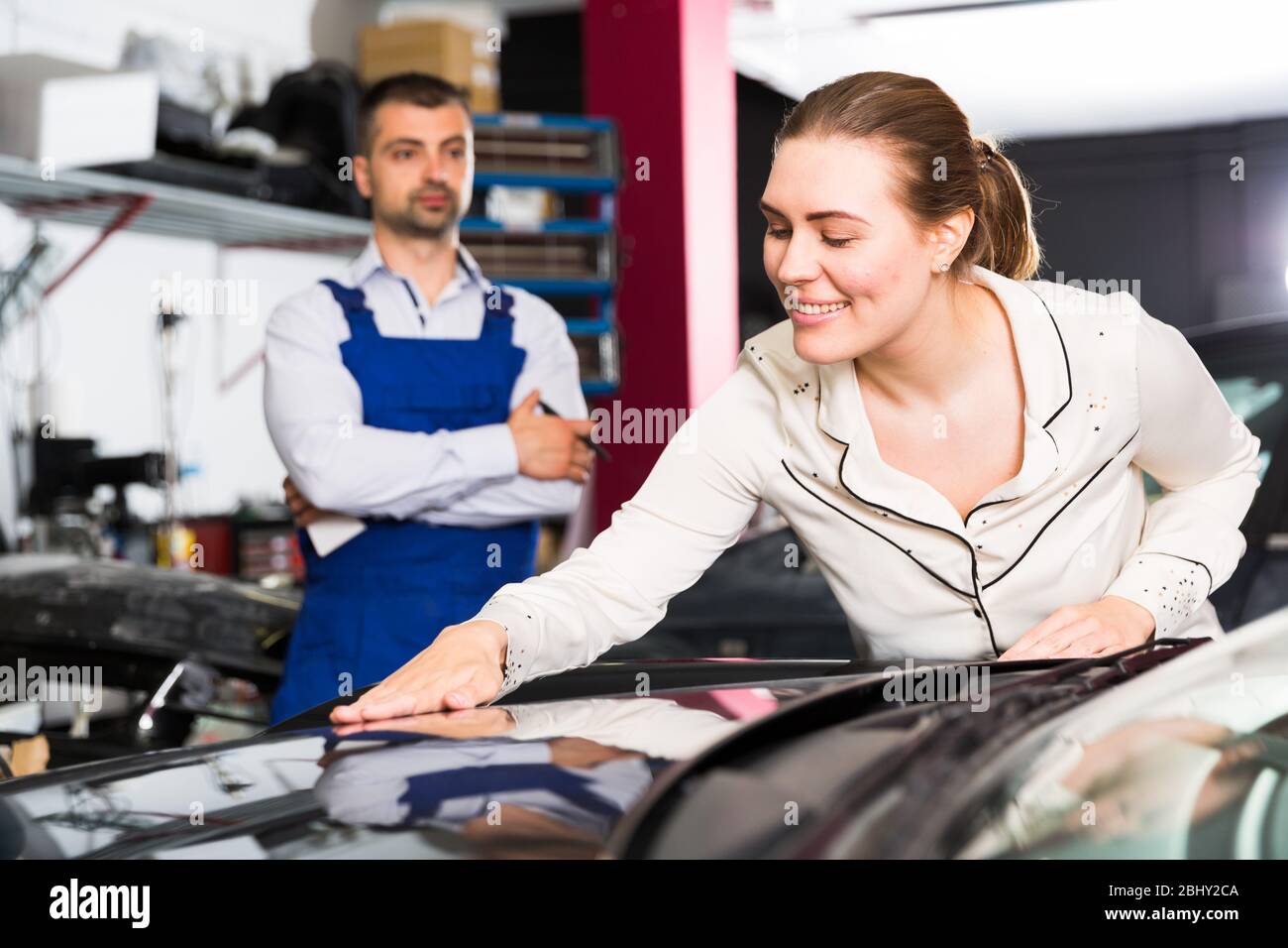 Happy young woman touching surface of repainted car body in auto repair shop Stock Photo