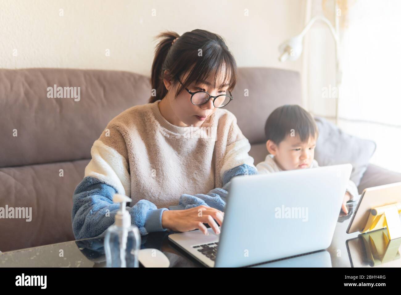 Young Asian mother working on a modern laptop from home with her child watching cartoon on a tablet during the coronavirus pandemic lockdown social di Stock Photo