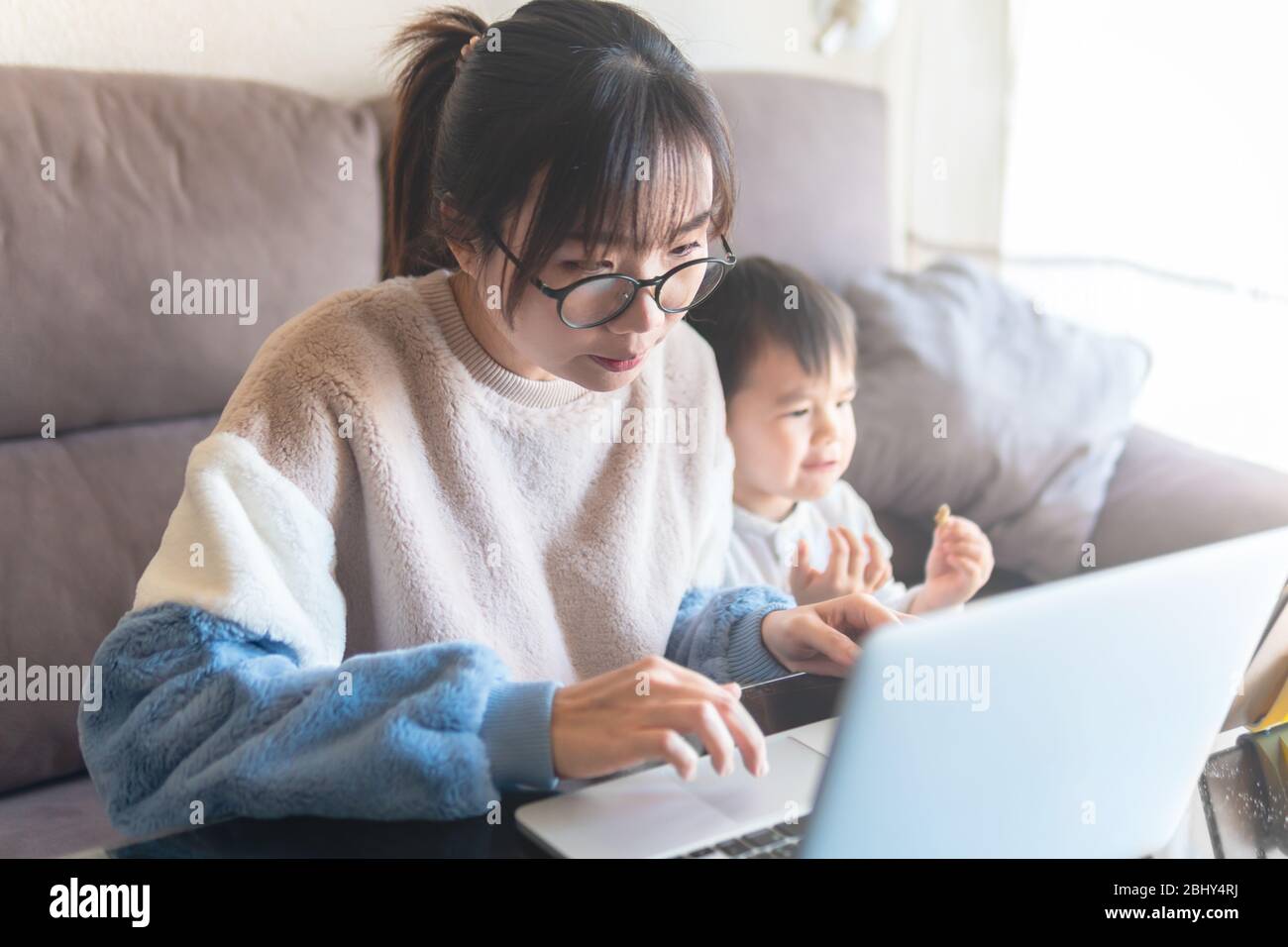 Young Asian mother working from home on computer. Kid watching cartoon on a tablet while mom work on laptop during the coronavirus pandemic lockdown Stock Photo