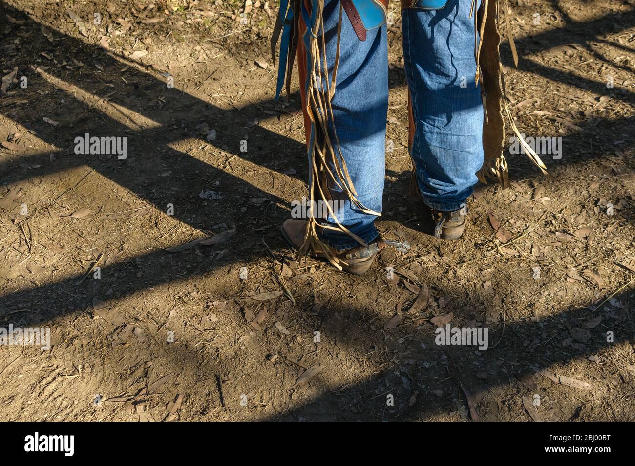 Cowboy legs with jeans, chaps, boots and spurs in the raking afternoon sun with his legs and rodeo rail shadows at Mareeba in Australia. Stock Photo