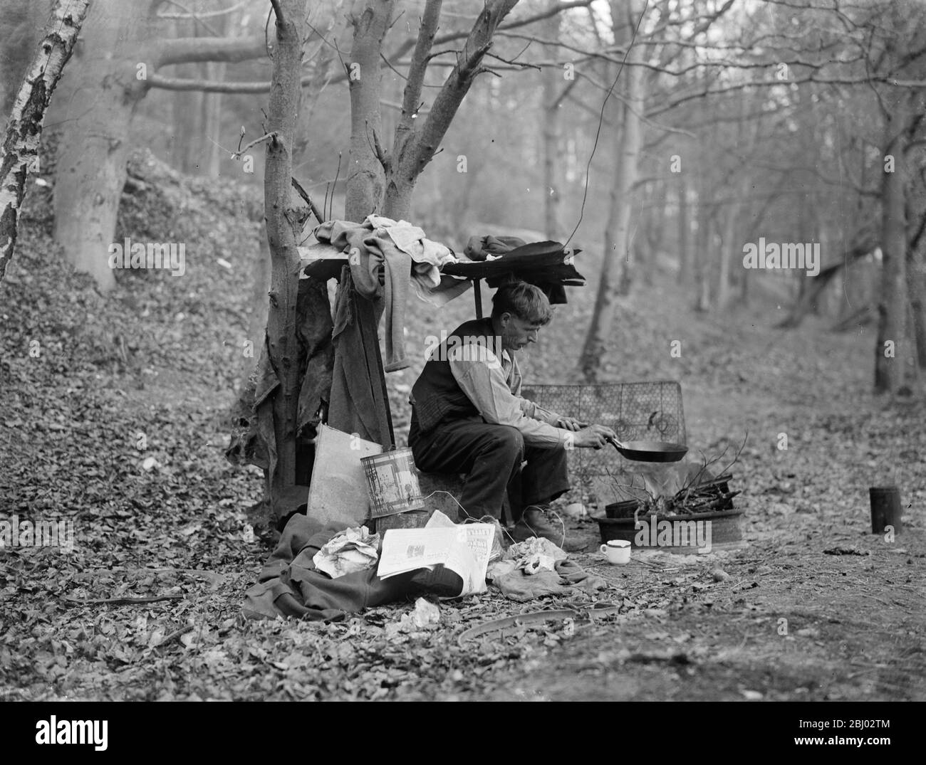 Tramp at home in woods cooking food over a camp fire . - 1935 Stock Photo
