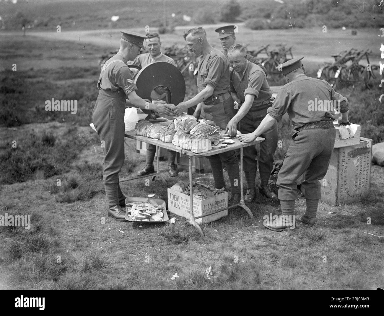 Behind The Orderly ' s Back . - Photo shows : While the orderlies are busily cutting supplies for troops of the 2nd Division Marlborough Lines , one of the men neatly helps himself to a slice of bread . The troops , now marching on manoeuvres through Hampshire and Sussex , camped on Frensham Common near Aldershot . - 28 Jul 1936 Stock Photo
