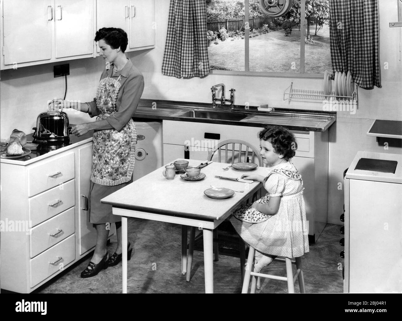 A idyllic view of a 1950s kitchen. The Mother puts some toast on while her daughter waits for breakfast. Stock Photo