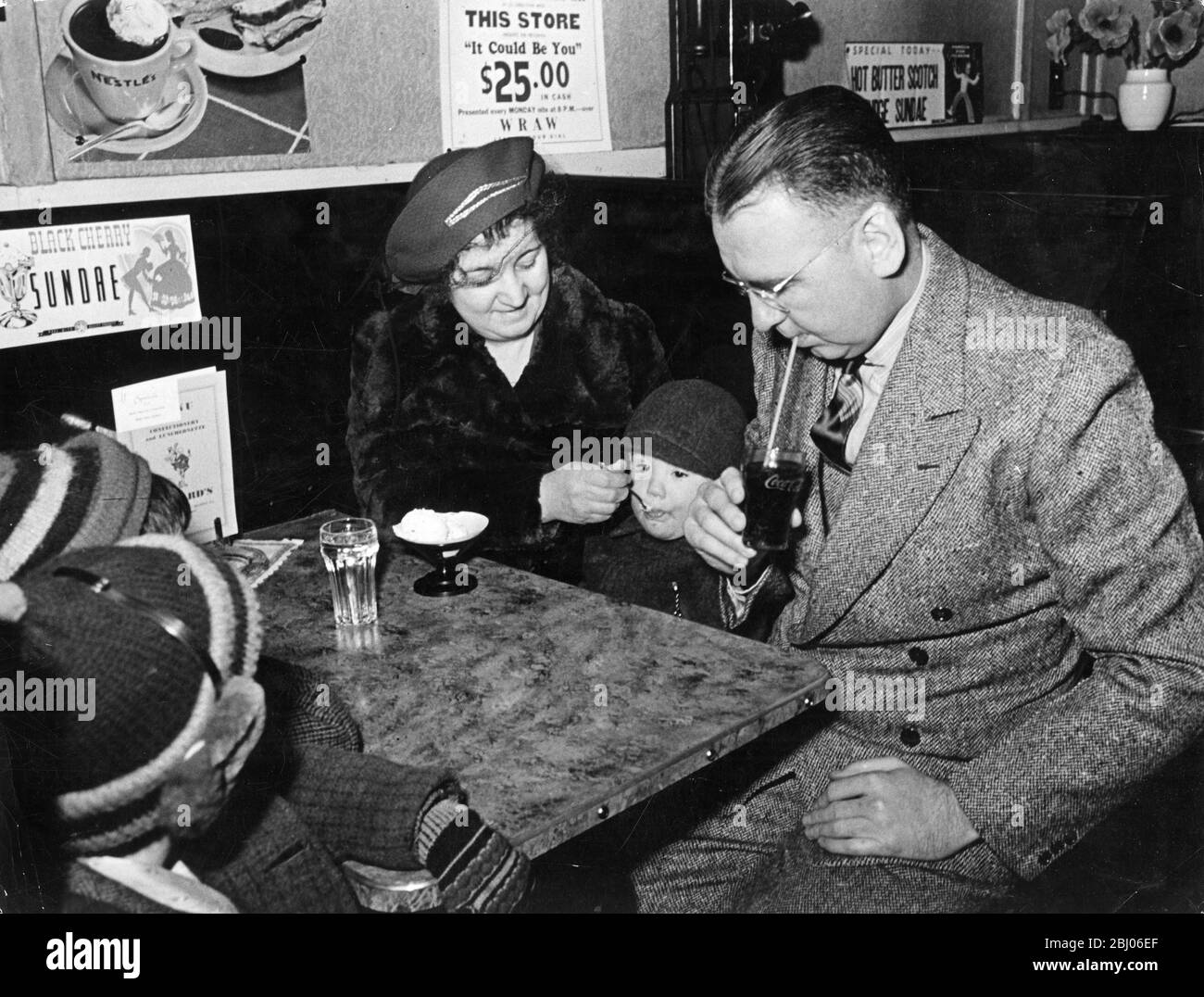 After their visit to a cinema Dr Corrigan and his family fo for some refreshments to a drug store. Mr Corrigan is sipping a Coca Cola while his wife is feeding ice cream to one of the children. Stock Photo