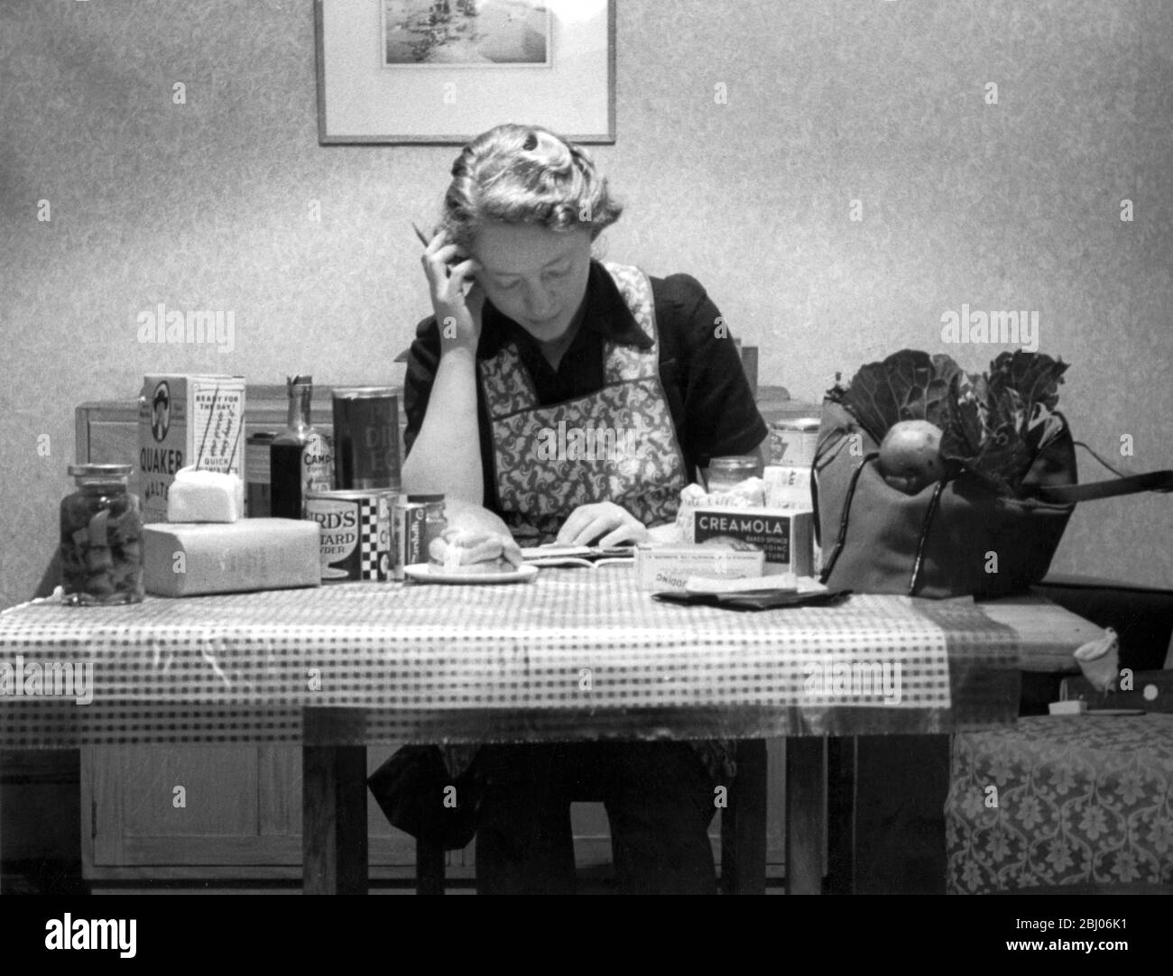 Housewife calculating the weekly household budget, with the food shopping laid out on the table. 1950's Stock Photo