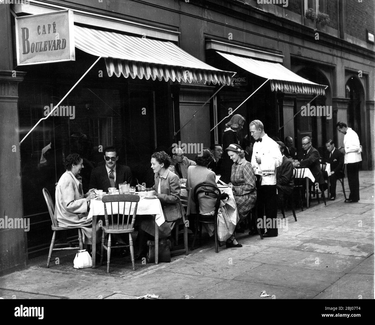 The unbeing usual scene outside the Cafe Boulevard in Shepherd's Place , Mayfair , off Upper Brook Street , London , shows a touch of continental living with diners outside on the pavement being attended to with waiter service . - 15 August 1951 Stock Photo