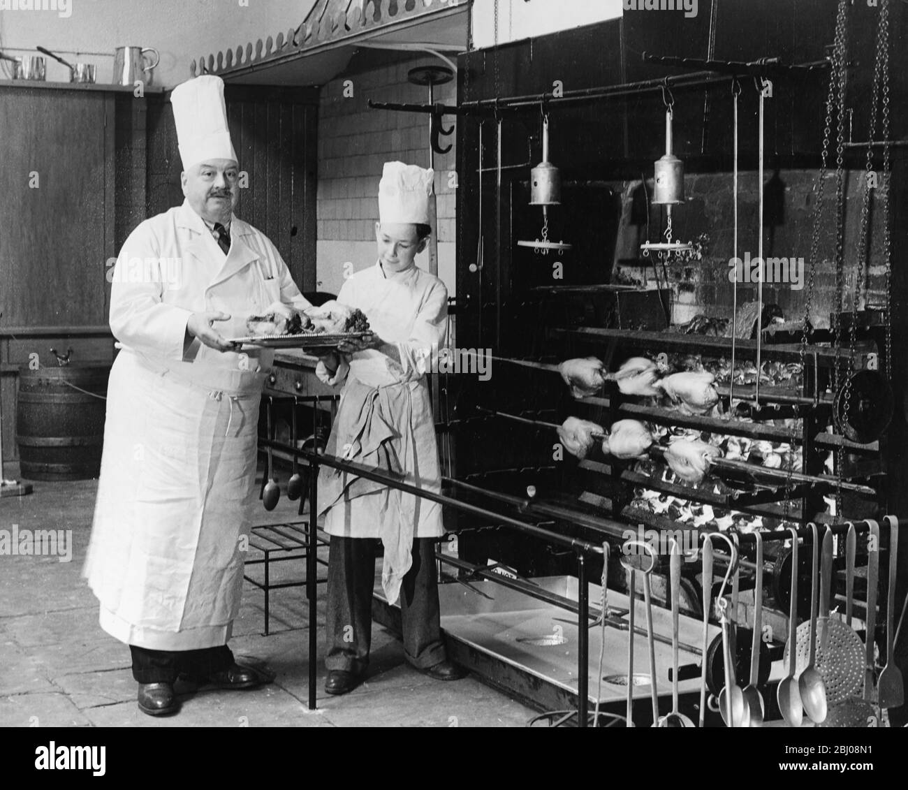 A.H. Cadier, senior chef at the Brighton Pavilion is handed fake chickens by junior chef Lloyd Payne to demonstrate the operation of the huge open fireplaces of 1825 in the kitchens at the Royal Pavilion. Brighton, Sussex, England - 16 July 1951 - Stock Photo