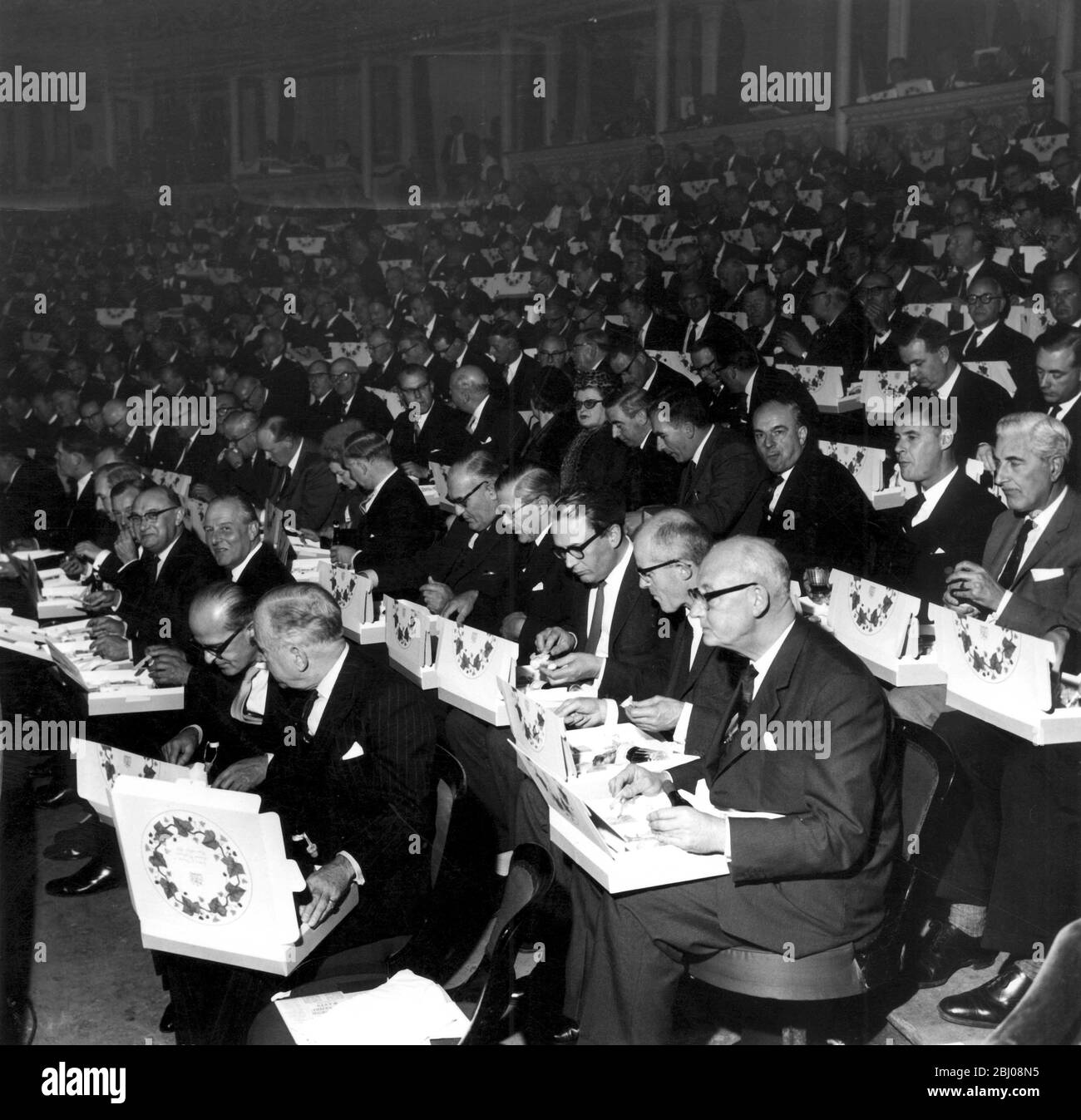 With luncheon boxes resting on their knees, some of the 5,000 company directors attending today's annual conference of the Institute of Directors in London's Royal Albert Hall, partake of an informal meal during a break in the proceedings. Each luncheon box contained a plastic knife and fork, mousse de foi gras, grilled lamb cutlet, roast chicken, stilton cheese, a bottle of red wine, a plastic glass, (cup or spoon), three cigarettes, a box of matches and a toothpick and tumbler. The Duke of Edinburgh attended this morning's session of the meeting, but did not stay for lunch. - 31st October 19 Stock Photo