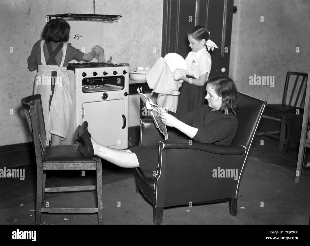 A Family of Sixteen - Mrs O Hara takes a break by putting her feet up and reads after the evening meal while her daughters clear up by doing the washing of the dishes 1949 Stock Photo