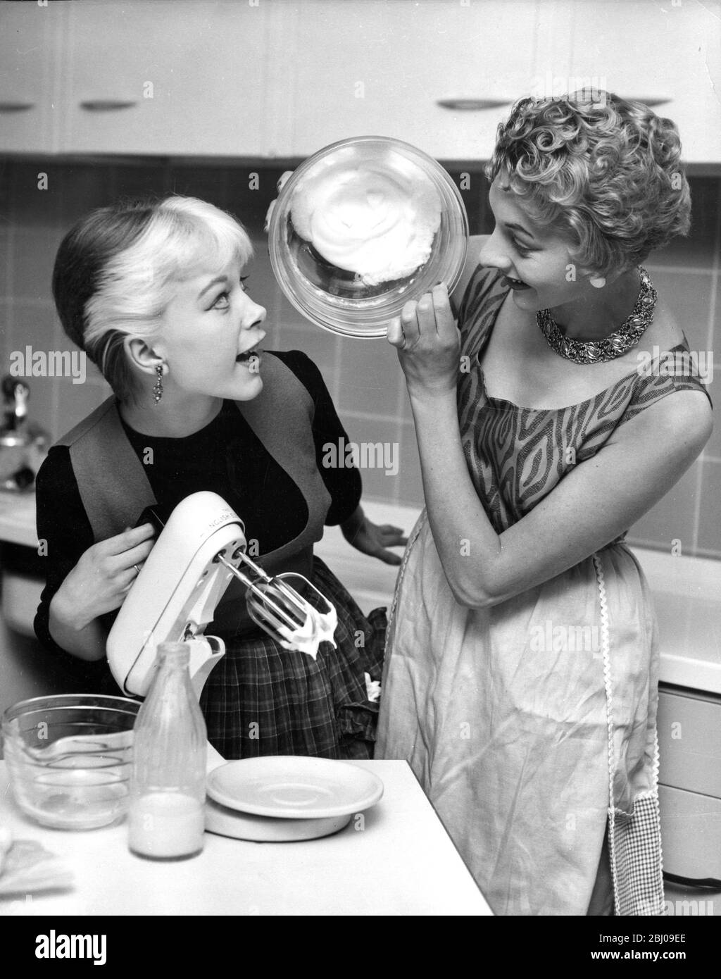 Model Zoe Newton , 18 , the  Milk Girl  whose smiling face on advertisements has made her familiar to millions is seen here (left) getting some culinary tips from Children's Television cookery expert Jacqueline Rose . Jacky is showing her how the whipped-up egg whites stay in a upturned bowl when the eggs have been beaten to the correct consistency . - 26 August 1954 Stock Photo