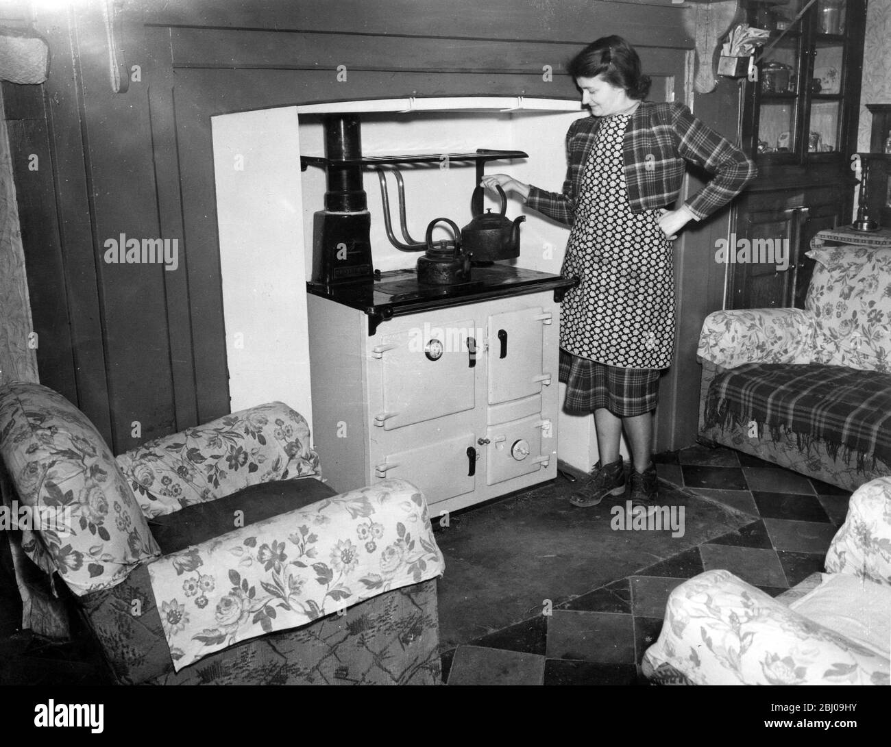 A woman boils to kettle for her cup of tea , in the main room of her cottage . The Rayburn range cooker also helps to keep the cottage warm and cosy , Kent , England . - 1930's Stock Photo