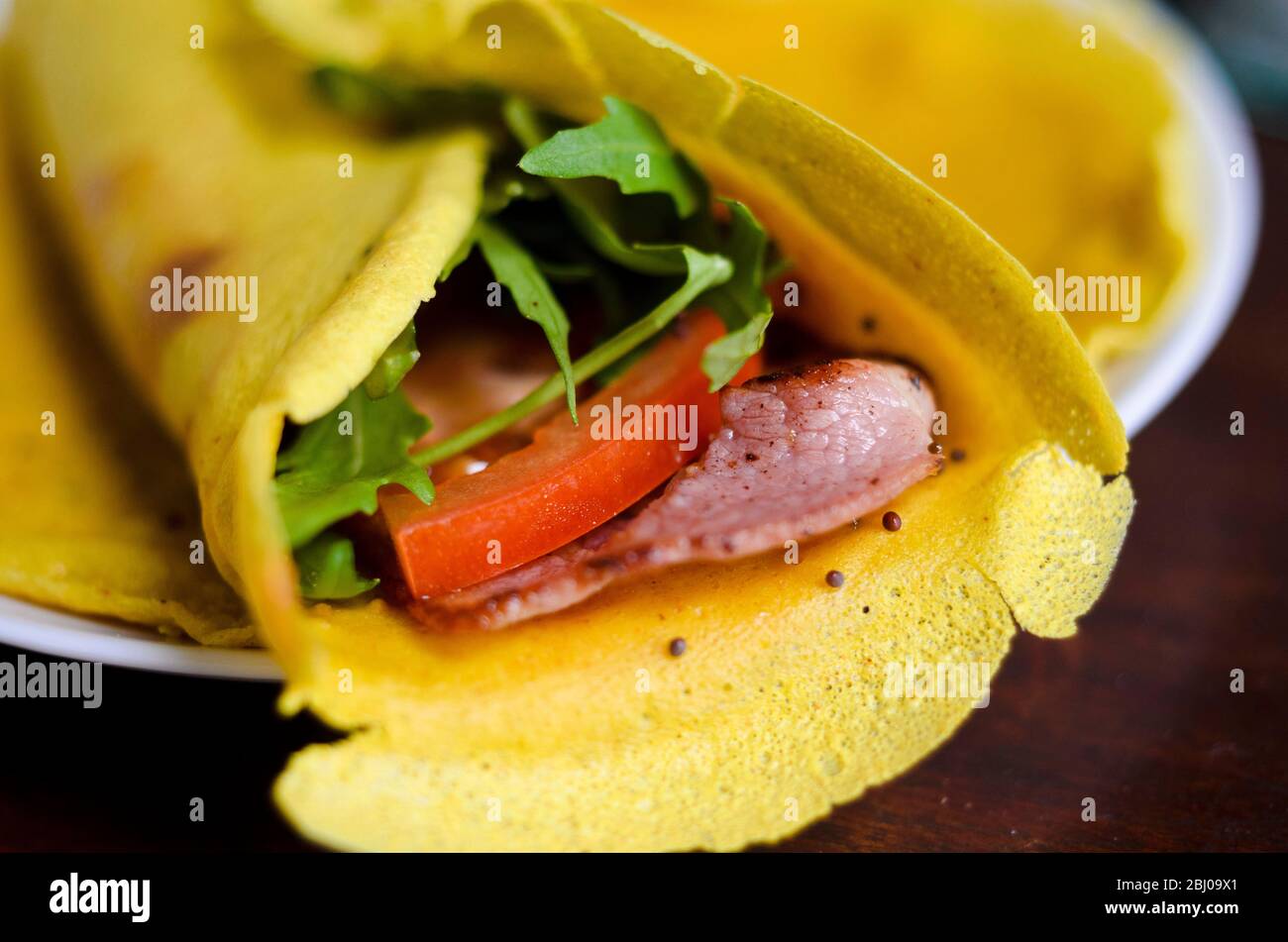 Pancake wrap made of gram (chickpea) flour, flavoured with turmeric, cumin and black onion seeds with a filling of rocket, tomato slices and a rasher Stock Photo
