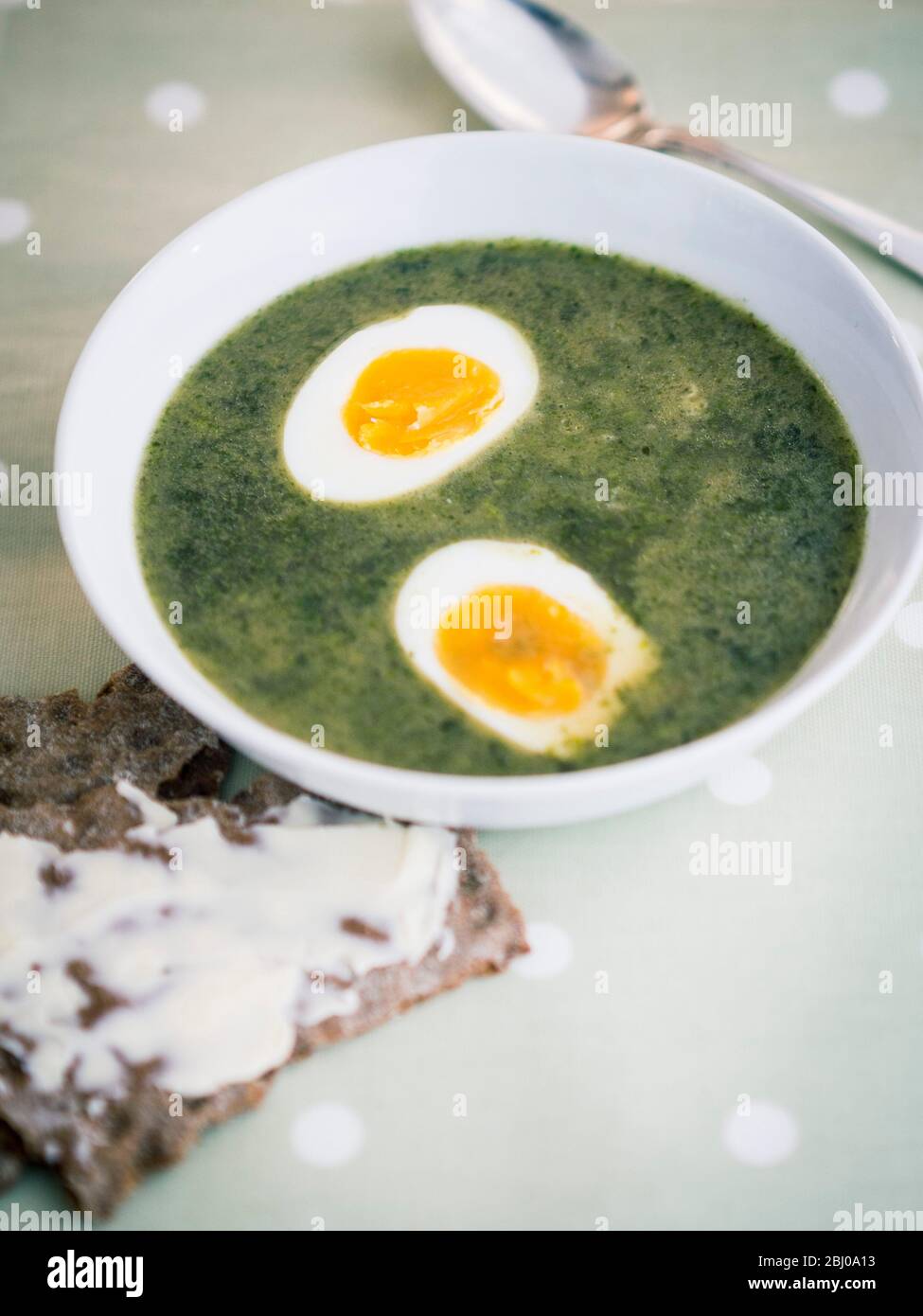 White bowl of Swedish nettle soup on striped cloth with silver spoon soup on spotted greeen cloth with Swedish knÌÄå ckebrÌÄå¦d (rye crispbread and bu Stock Photo
