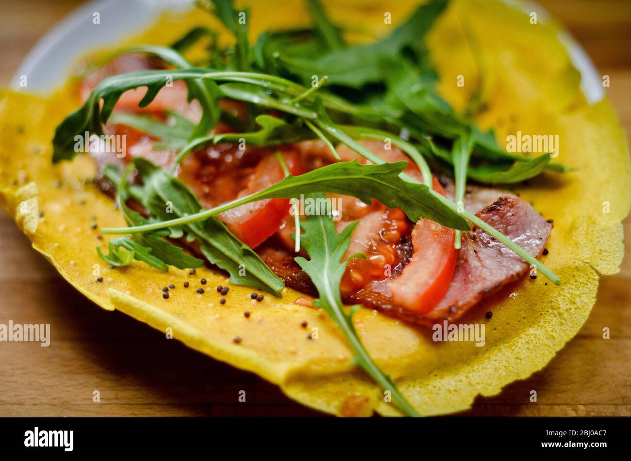 Pancake wrap made of gram (chickpea) flour, flavoured with turmeric, cumin and black onion seeds with a filling of rocket, tomato slices and a rasher Stock Photo