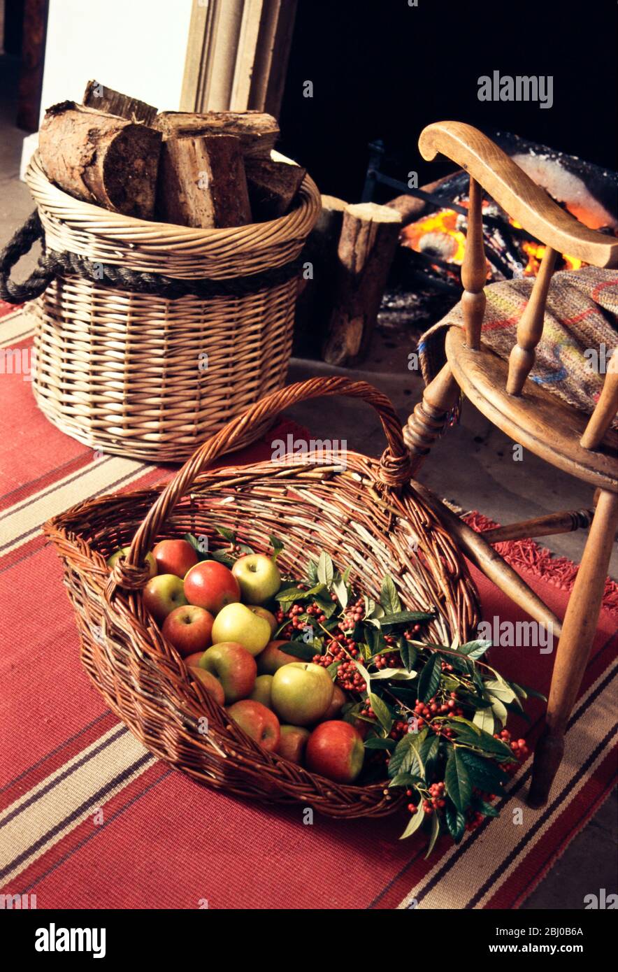 Big decorative basket of Cox's apples with sprays of ornamental berries by open fireplace with old fashioned chair and log basket - Stock Photo