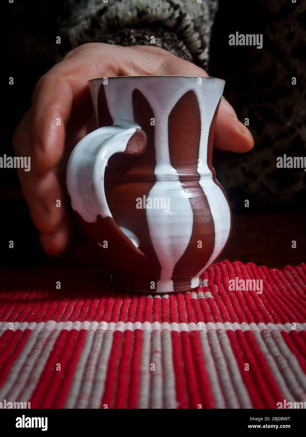 Picking up a cup of coffee or tea in a stripey pottery mug on a striped tablecloth - Stock Photo