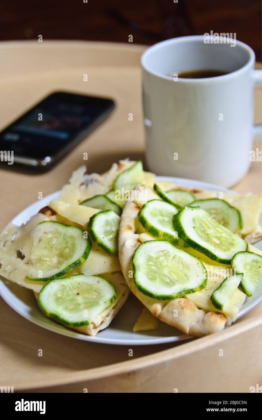 Snack of Scandinavian flatbread, with thinly sliced Swedish cheese and cucumber slices, sprinkled wih herb salt, with cup of black coffee and iphone o Stock Photo