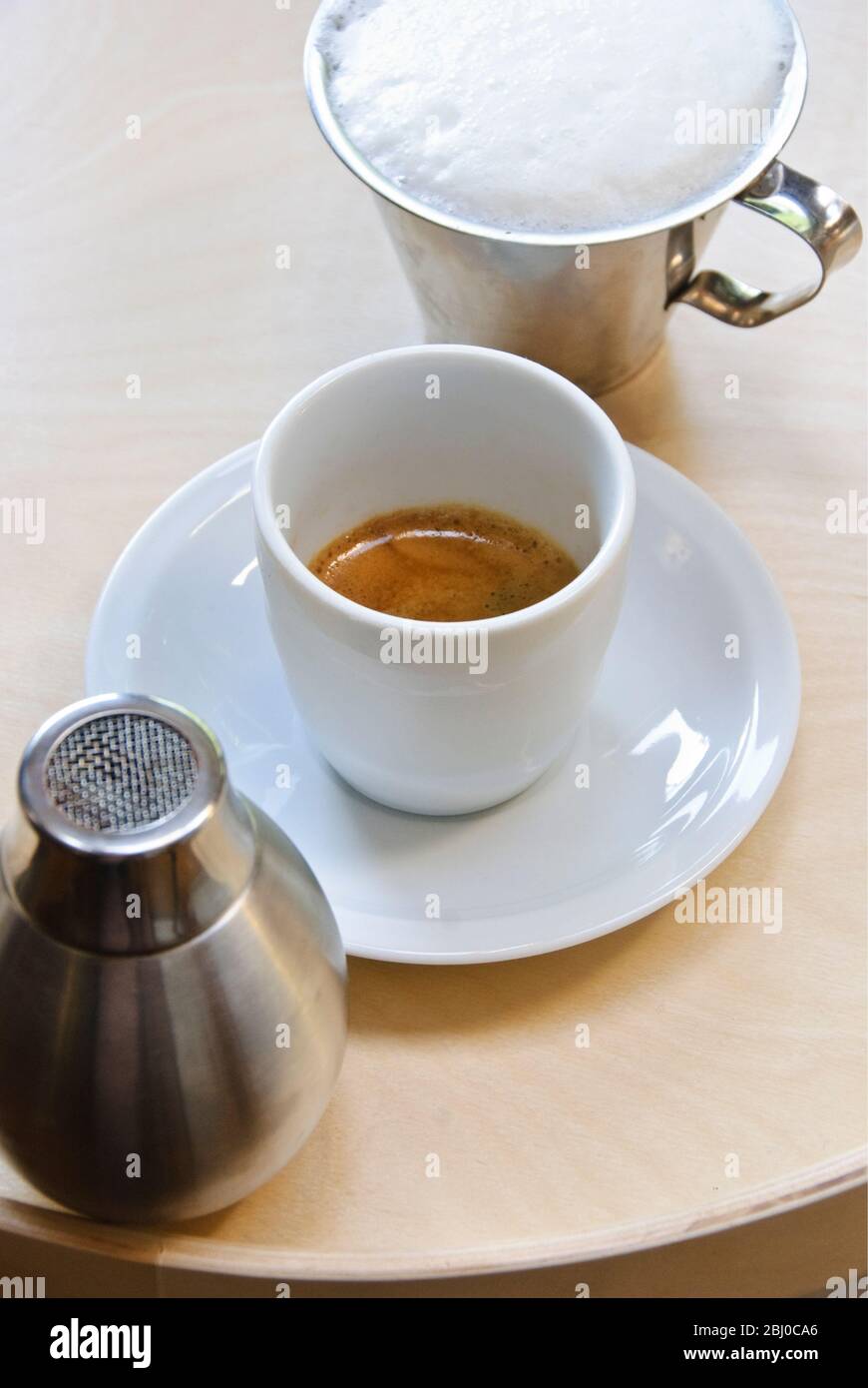 White porcelain cup of freshly brewed espresso coffee showing 'crema' with frothed milk in jug and cocoa shaker - about to turn the espresso into capp Stock Photo