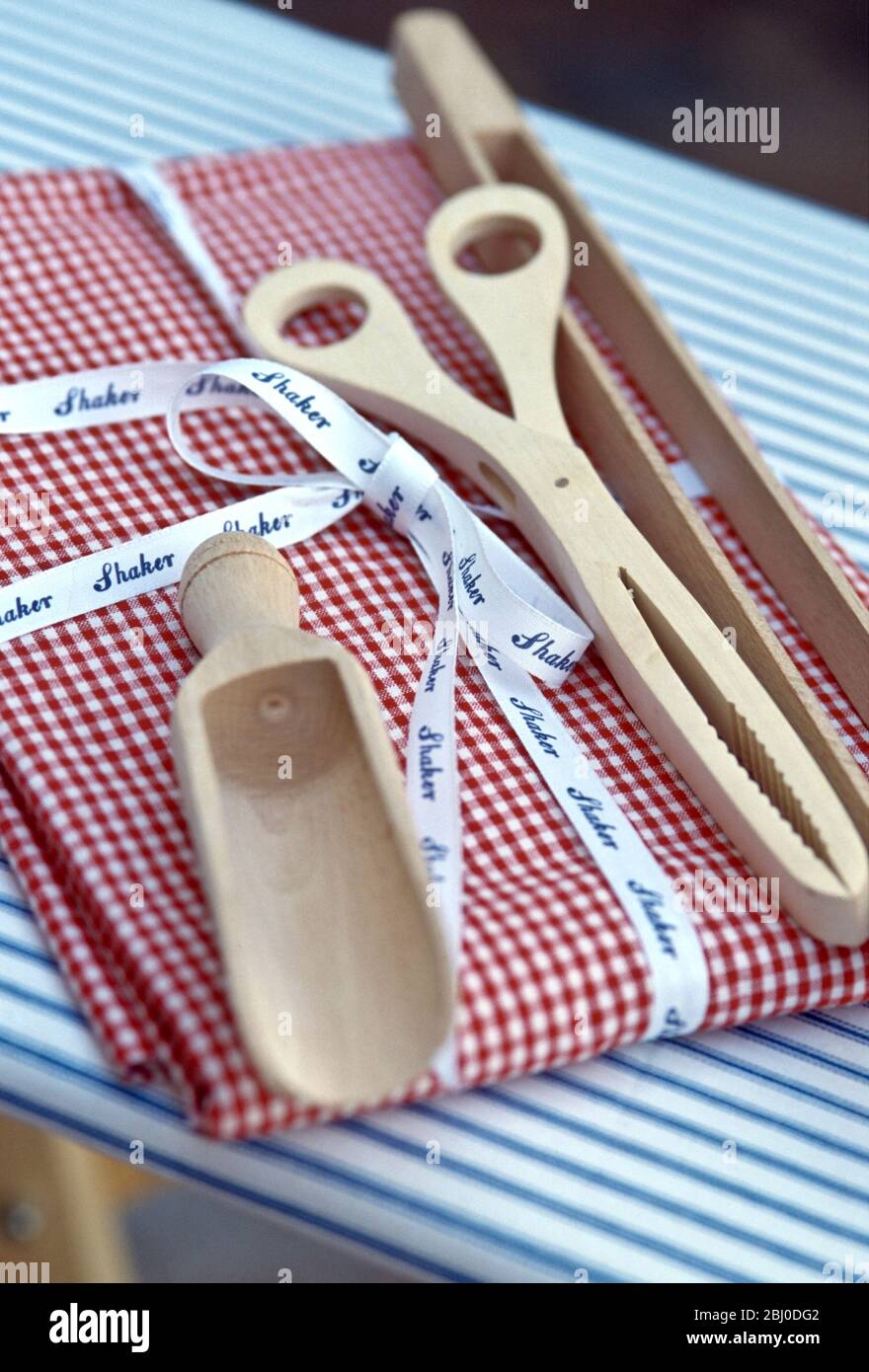 Wooden laundry tongs and detergent scoop on folded red gingham cloth on blue and white striped ironing board, with printed Shaker ribbon - Stock Photo