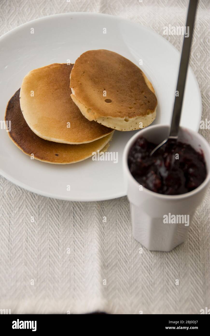 Three Scotch pancakes on a white plate with small pot of raspberry jam - Stock Photo