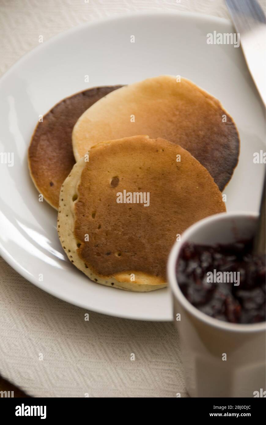 Three Scotch pancakes on a white plate with small pot of raspberry jam - Stock Photo