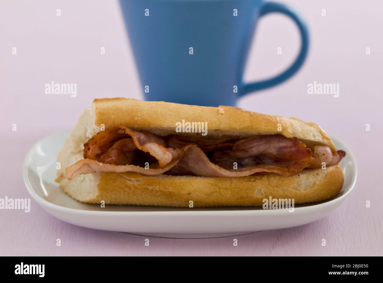 Streaky bacon in white baguette with blue mug of black coffee - Stock Photo