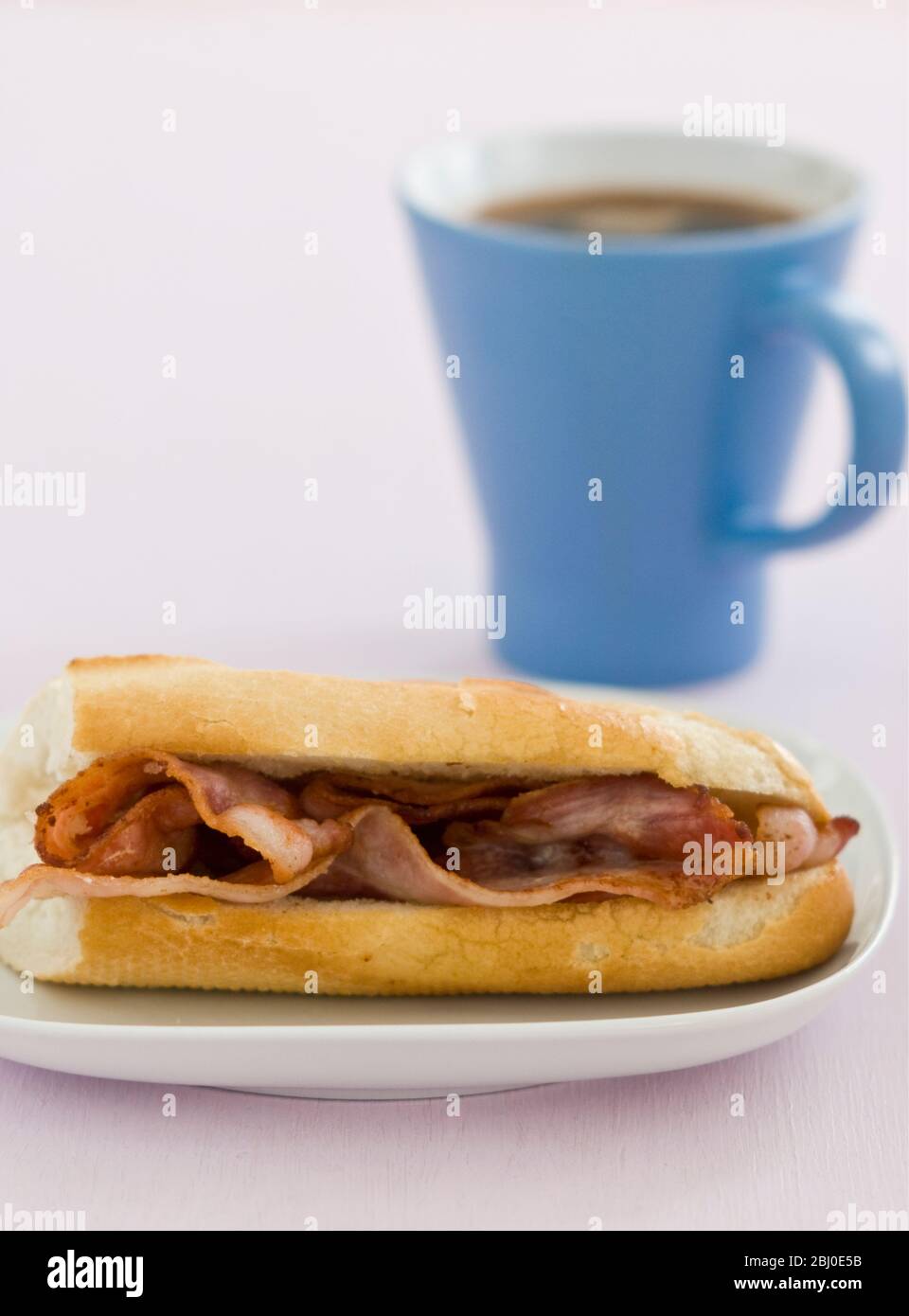 Streaky bacon in white baguette with blue mug of black coffee - Stock Photo