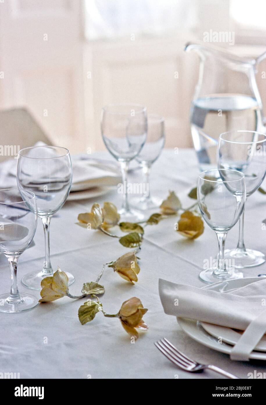 Paper roses on simple white tablecloth with plain crystal glasses and white china for an elegant party table setting - Stock Photo
