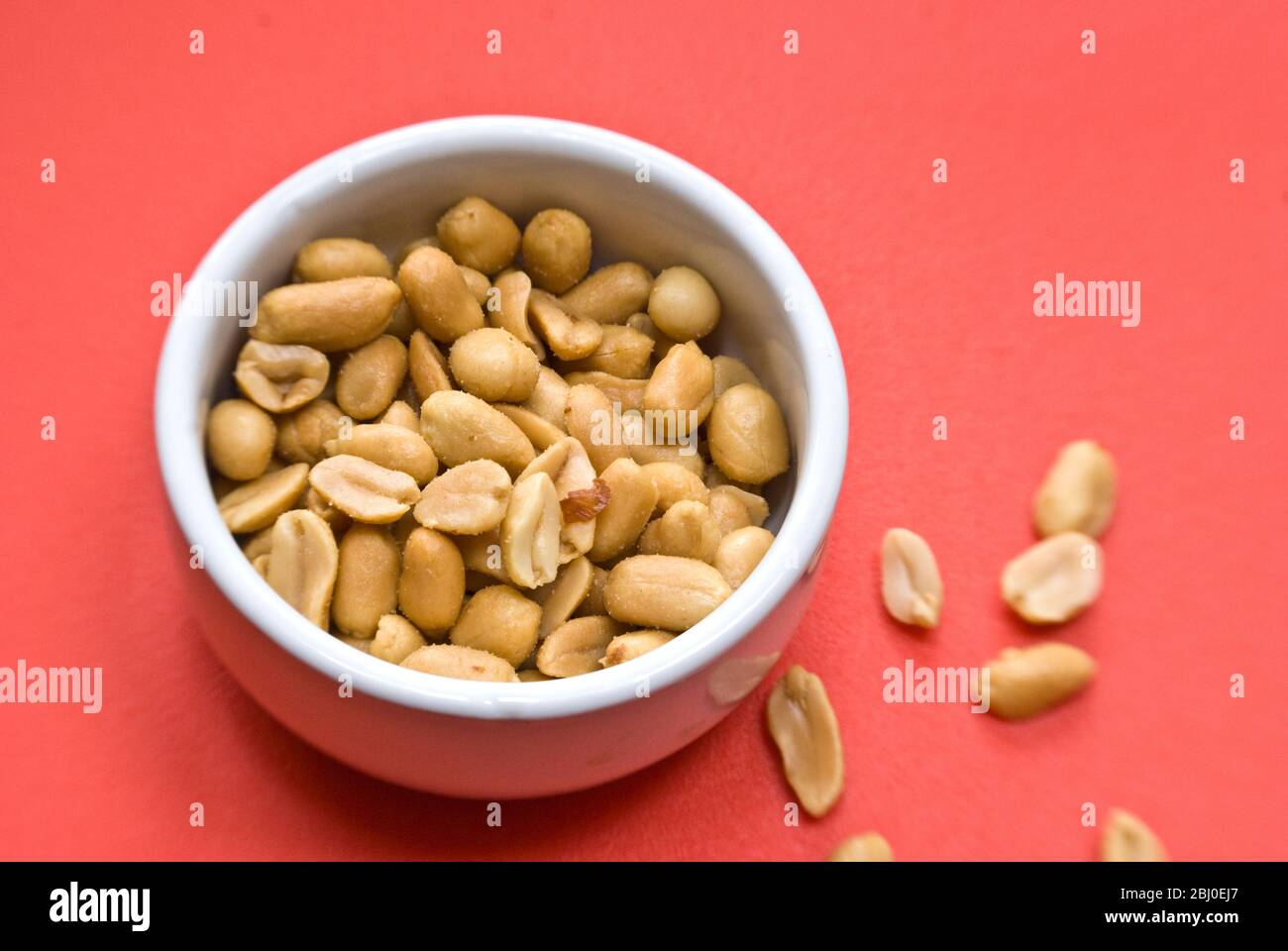 Small white bowl of salted, roasted peanuts on bright red background - Stock Photo