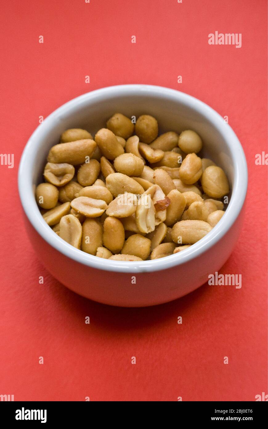 Small white bowl of salted, roasted peanuts on bright red background - Stock Photo