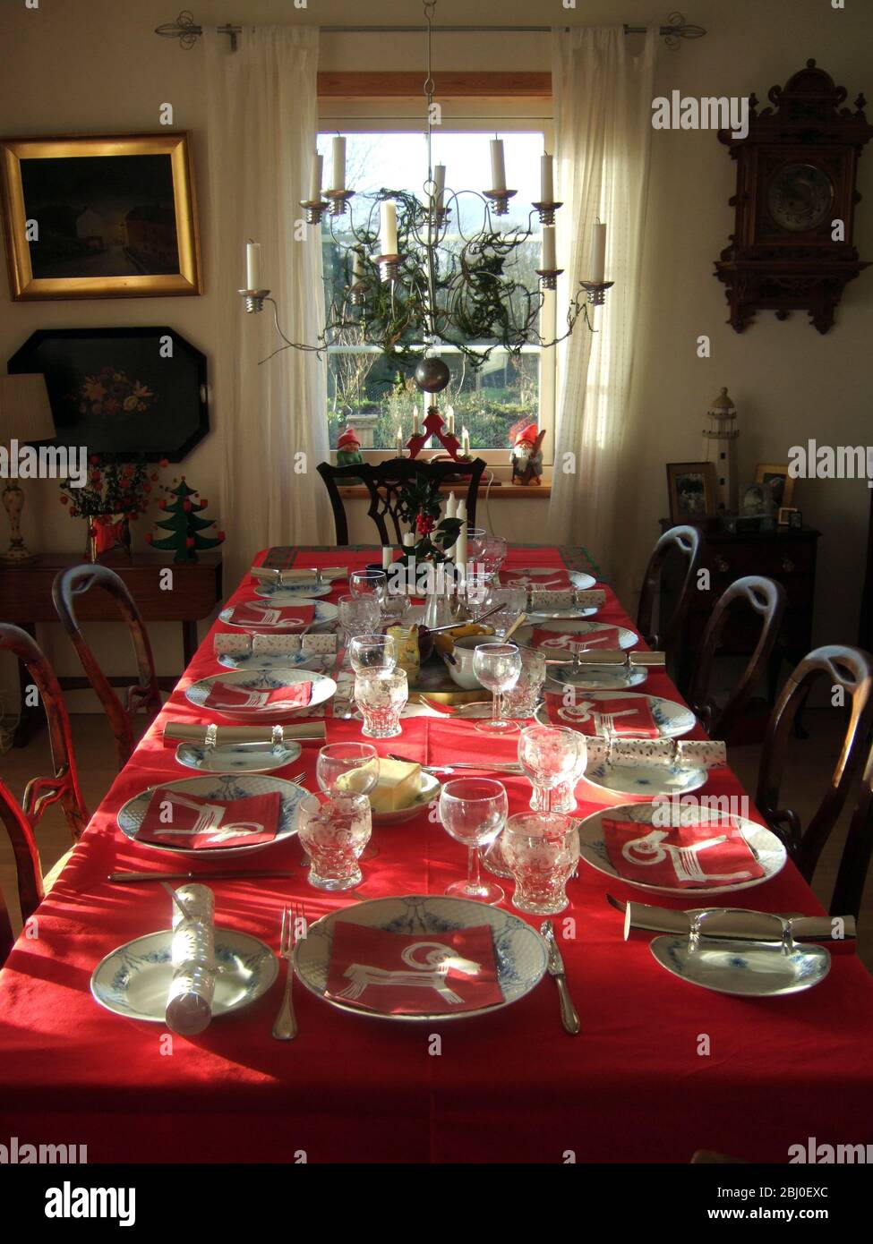 Sunlight shining in on interior with long table laid for Christmas meal with red tablecloth and best china, - Stock Photo