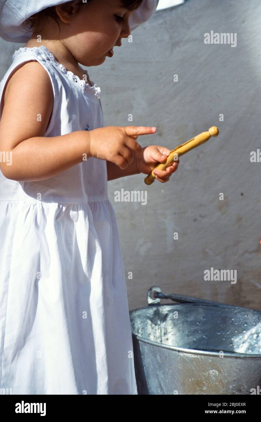 Little girl playing with a bucket of clear water and some clothes pegs while mother is hanging out the washing in sunny yard - Stock Photo