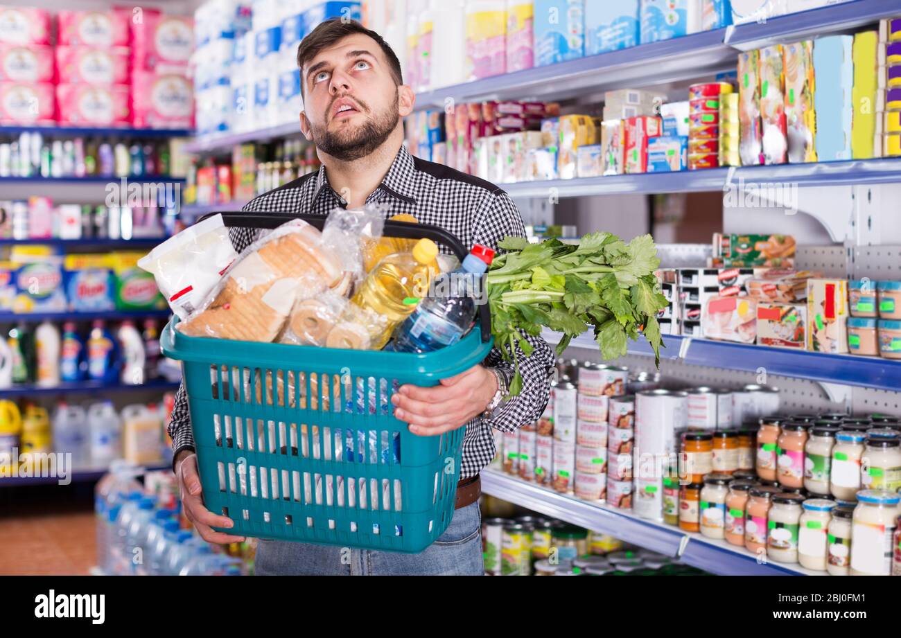 Tired young man with heavy shopping basket filled food products in supermarket Stock Photo