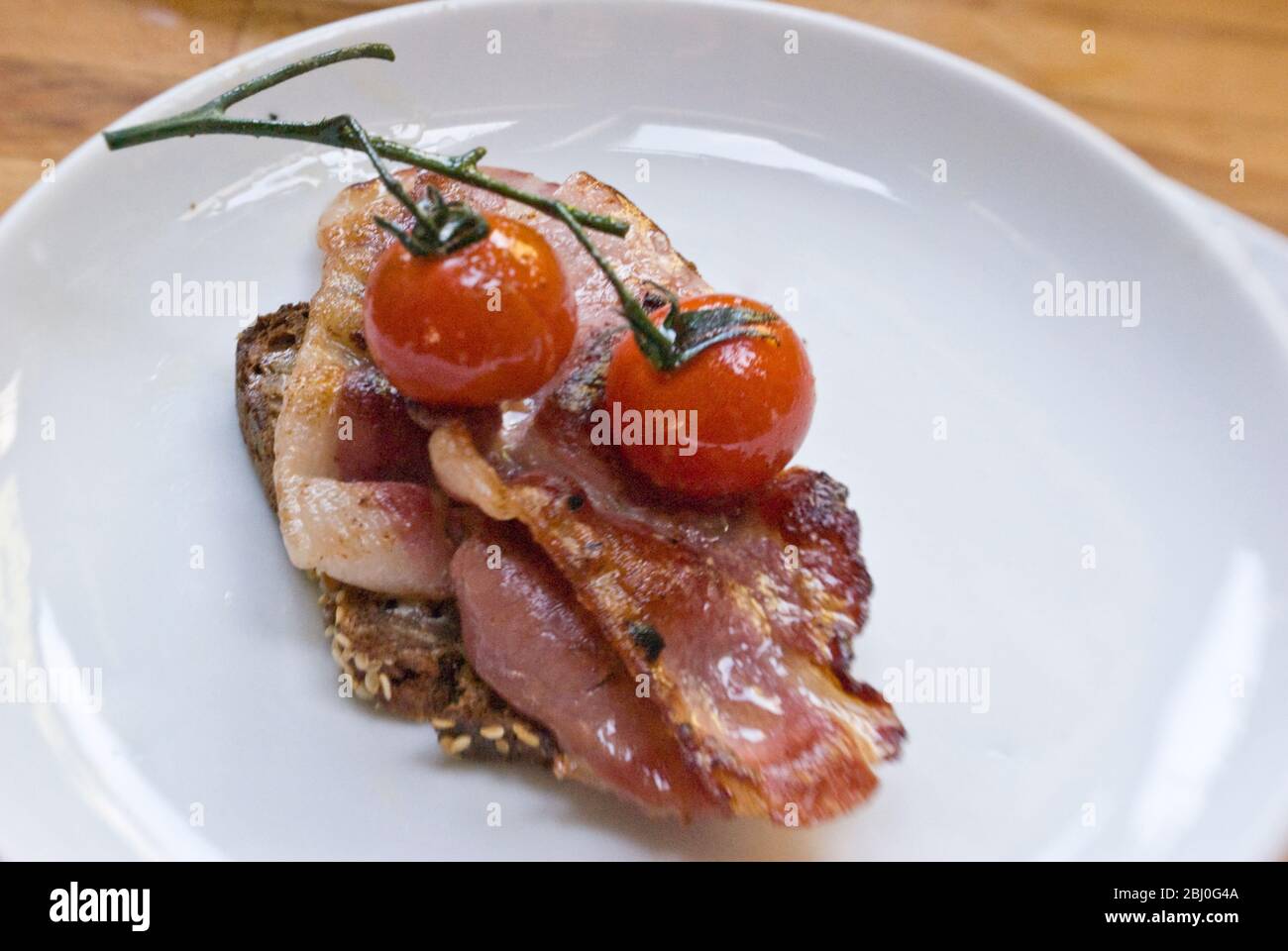 Breakfast dish of coarse wholemeal rye bread with organic bacon and griddled chery tomatoes on the vine - Stock Photo