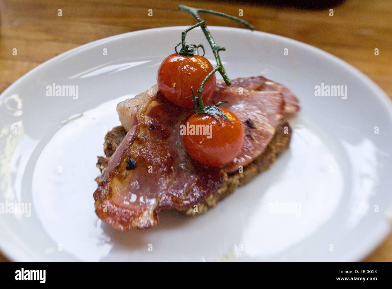 Breakfast dish of coarse wholemeal rye bread with organic bacon and griddled chery tomatoes on the vine - Stock Photo